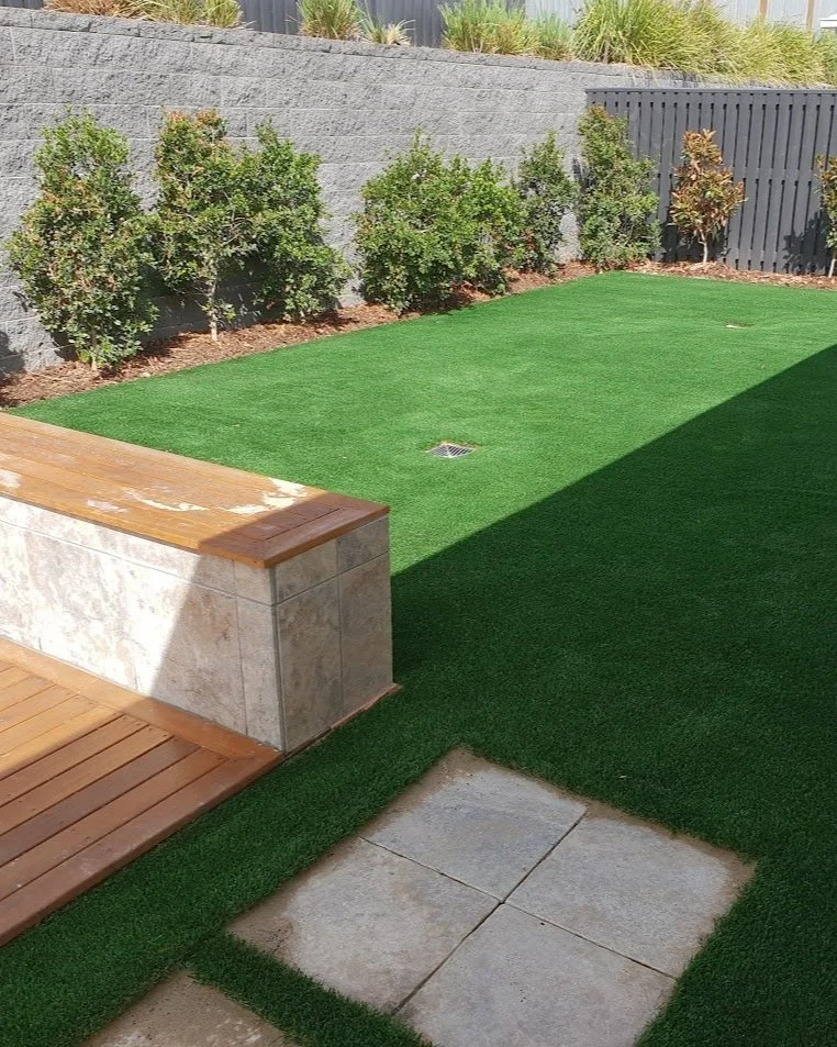 Backyard with artificial grass, small shrubs along stone wall, wooden deck, and paved tiles.