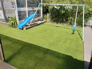 Backyard playground with slide, swing, green artificial turf, and plants.