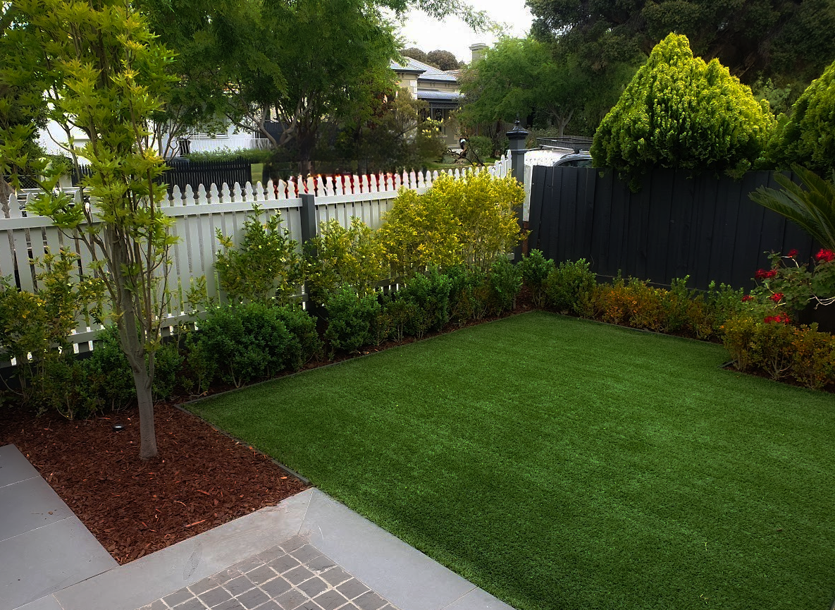 Backyard garden with green lawn and trees, surrounded by a white fence.