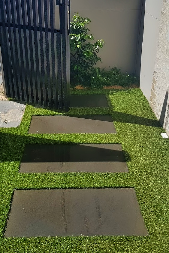 Outdoor pathway with rectangular stepping stones on artificial grass next to a black metal gate.