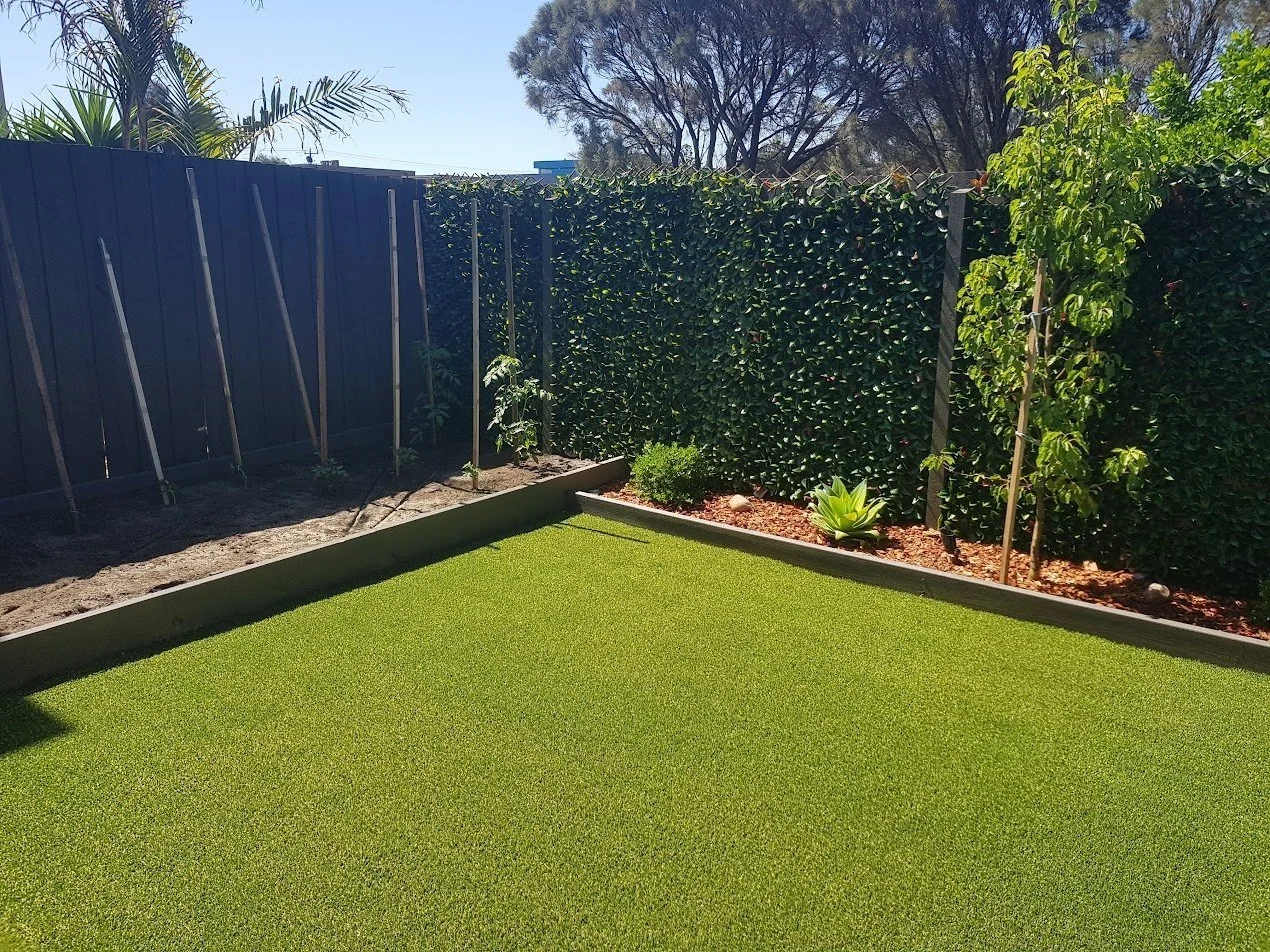 Backyard garden with artificial turf, wooden fence, plants, and a small garden bed with stakes.