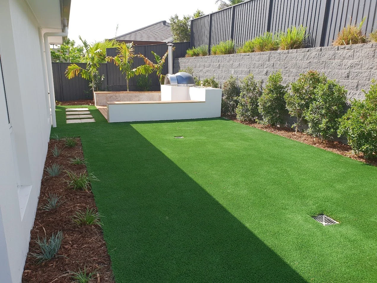 Backyard with artificial turf, small garden bed along the house, plants along concrete wall, and barbecue area.