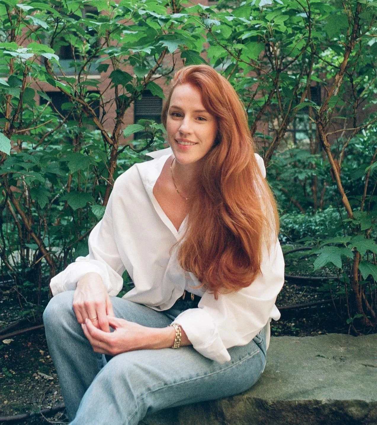 A woman with long red hair sitting outdoors on a rock, smiling, surrounded by green foliage, wearing a white shirt and light jeans.