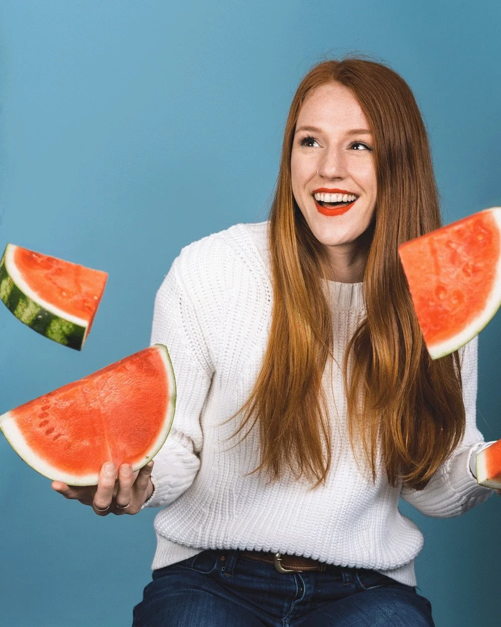 A woman with long red hair holding three slices of watermelon, smiling and looking to her right against a blue background.