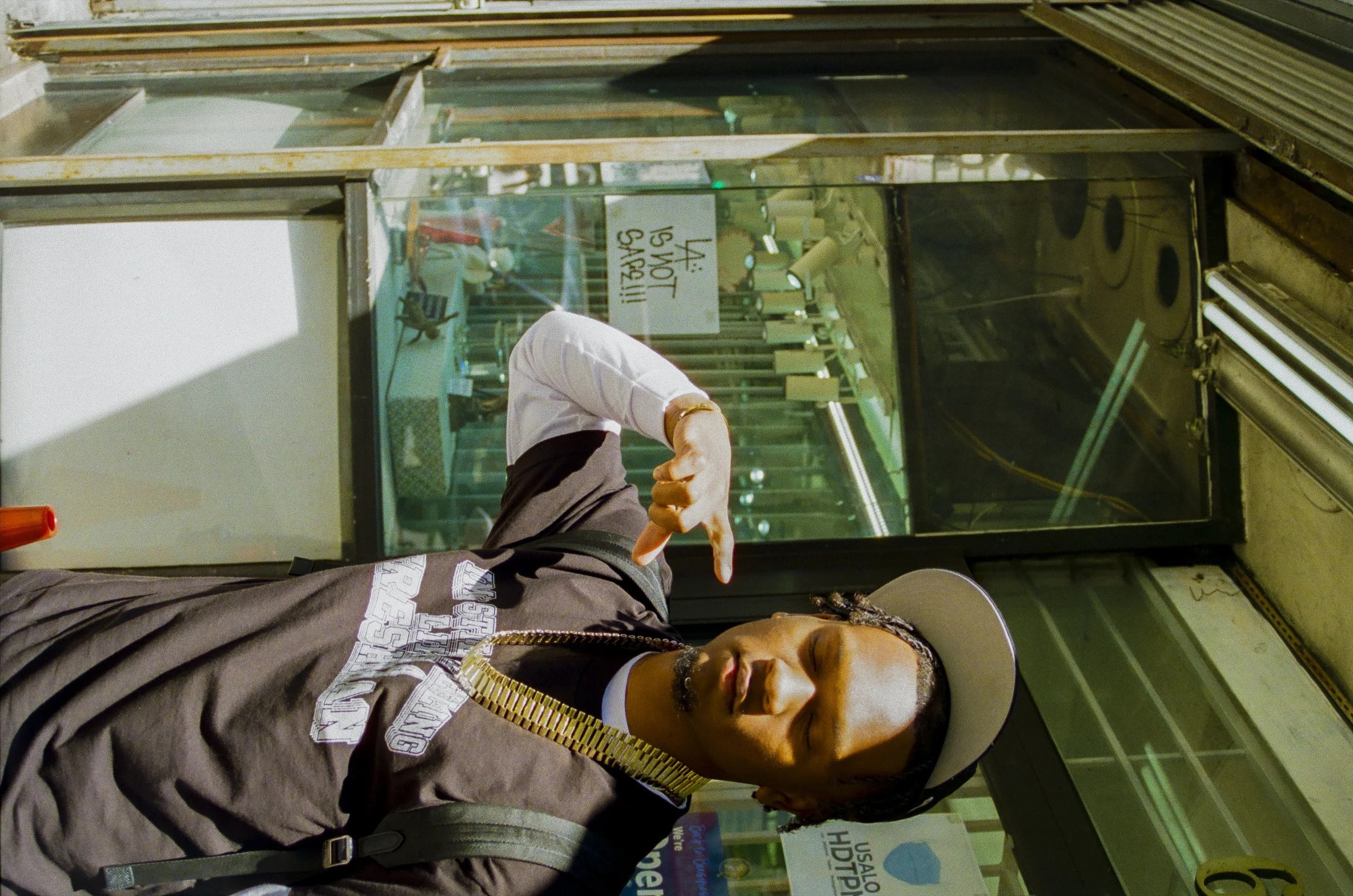 A person wearing a black t-shirt with white text and a gold chain stands in a shop, holding a piece of paper. Behind them, there is a glass display case with various items inside, and a woman is seen pointing at something inside the case. The backgro