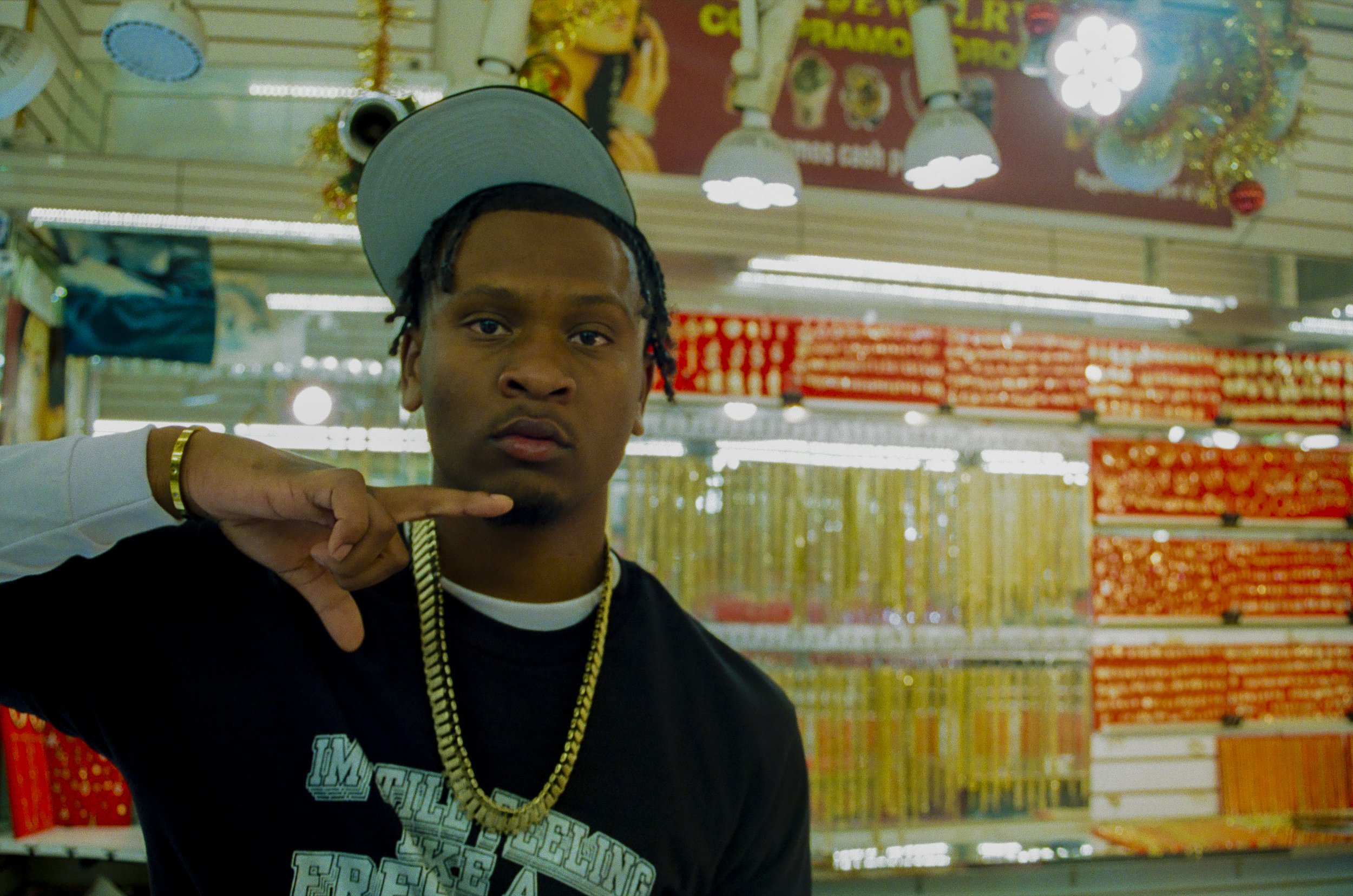 Young man with braided hair wearing a cap, gold chain, and black hoodie in a store aisle with colorful holiday decorations and shelves of red boxes in the background.