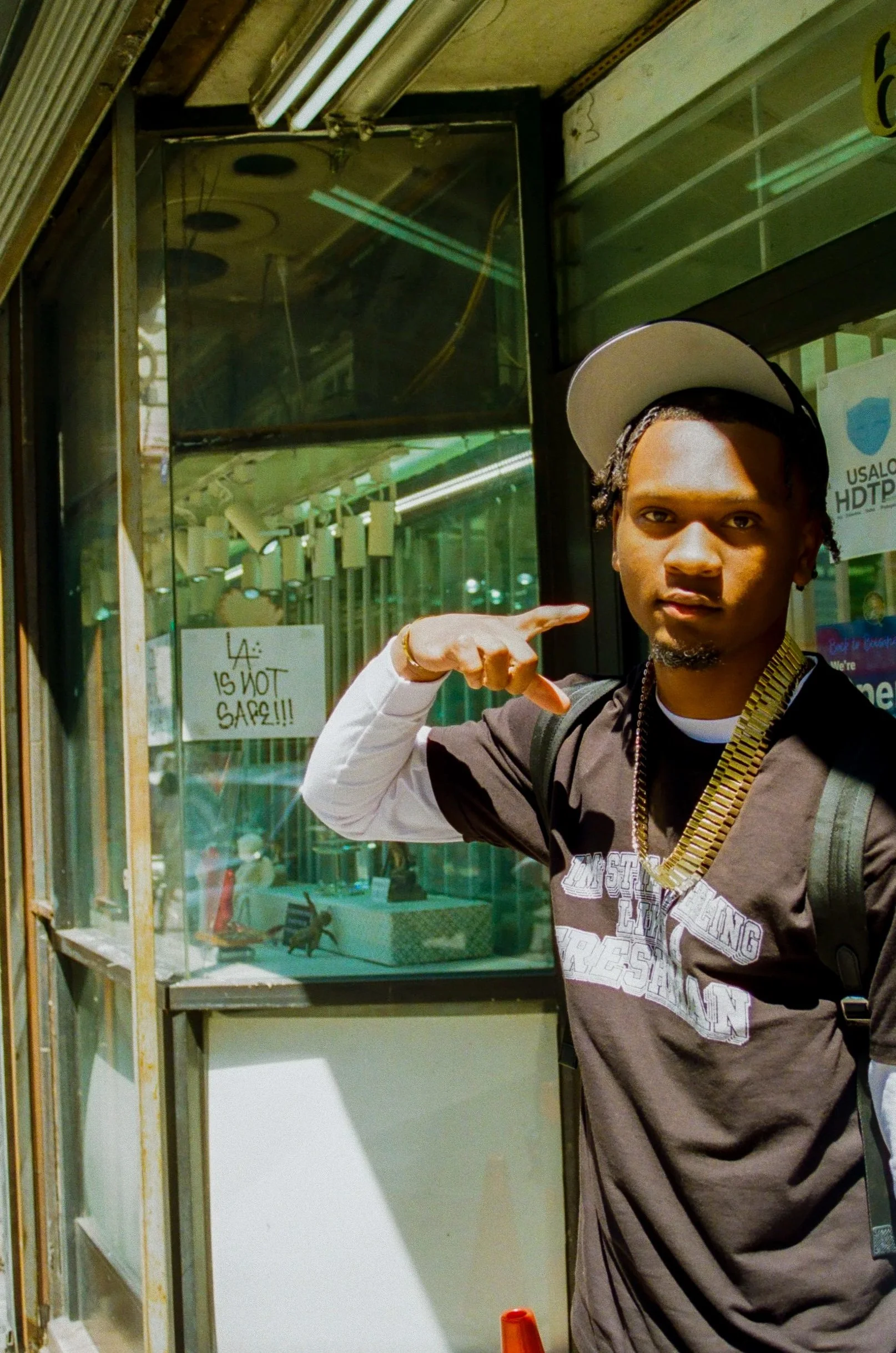 A young man with styled hair, wearing a gray T-shirt, pointing at a glass display case outside a store that has a sign reading 'LA IS NOT SAFE!!!' with a background of store windows and signs.