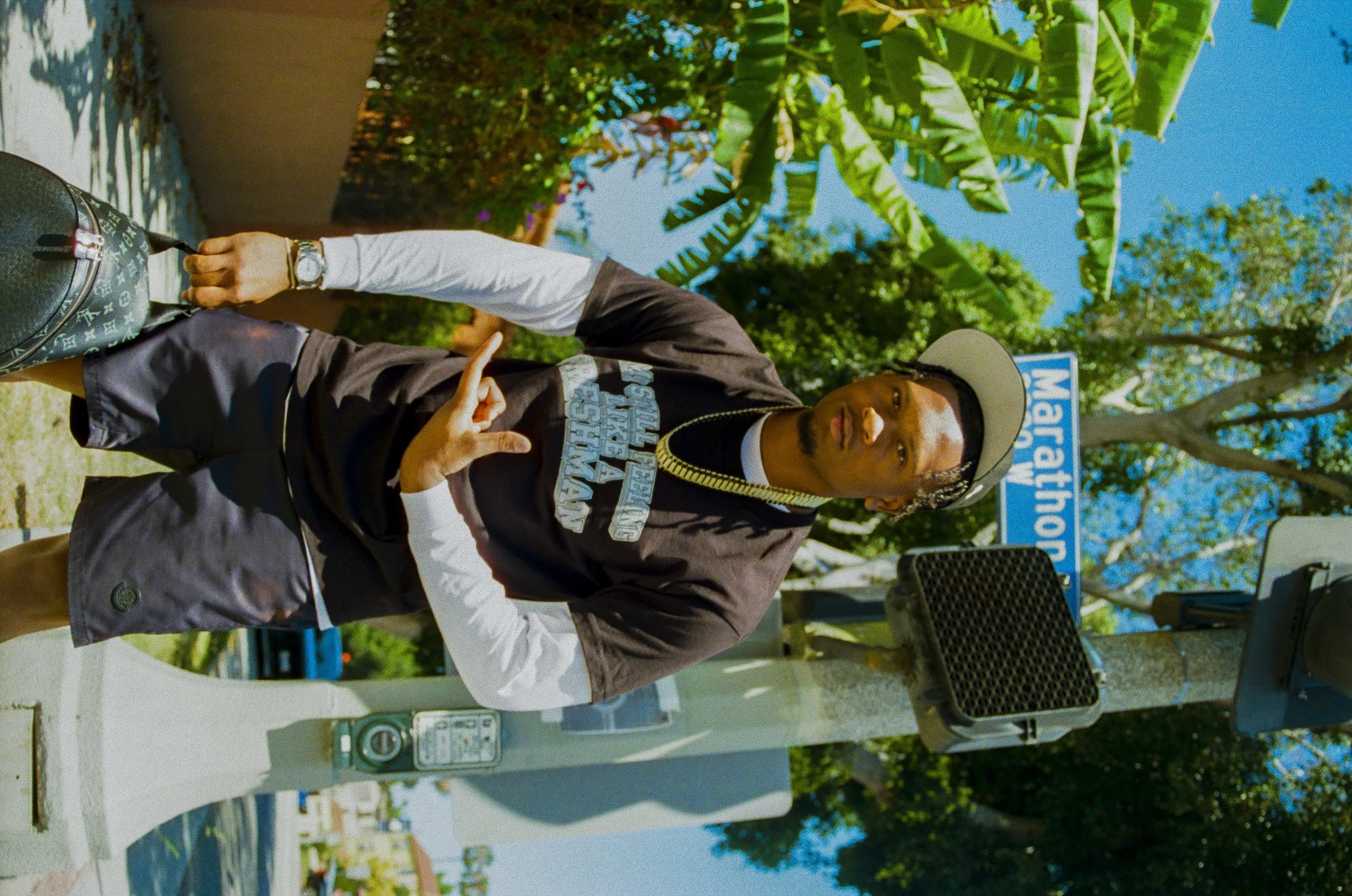 A man standing outdoors next to a parking meter, making a peace sign with his right hand, wearing a hat, jacket, and wristwatch, with trees and a blue sky in the background.