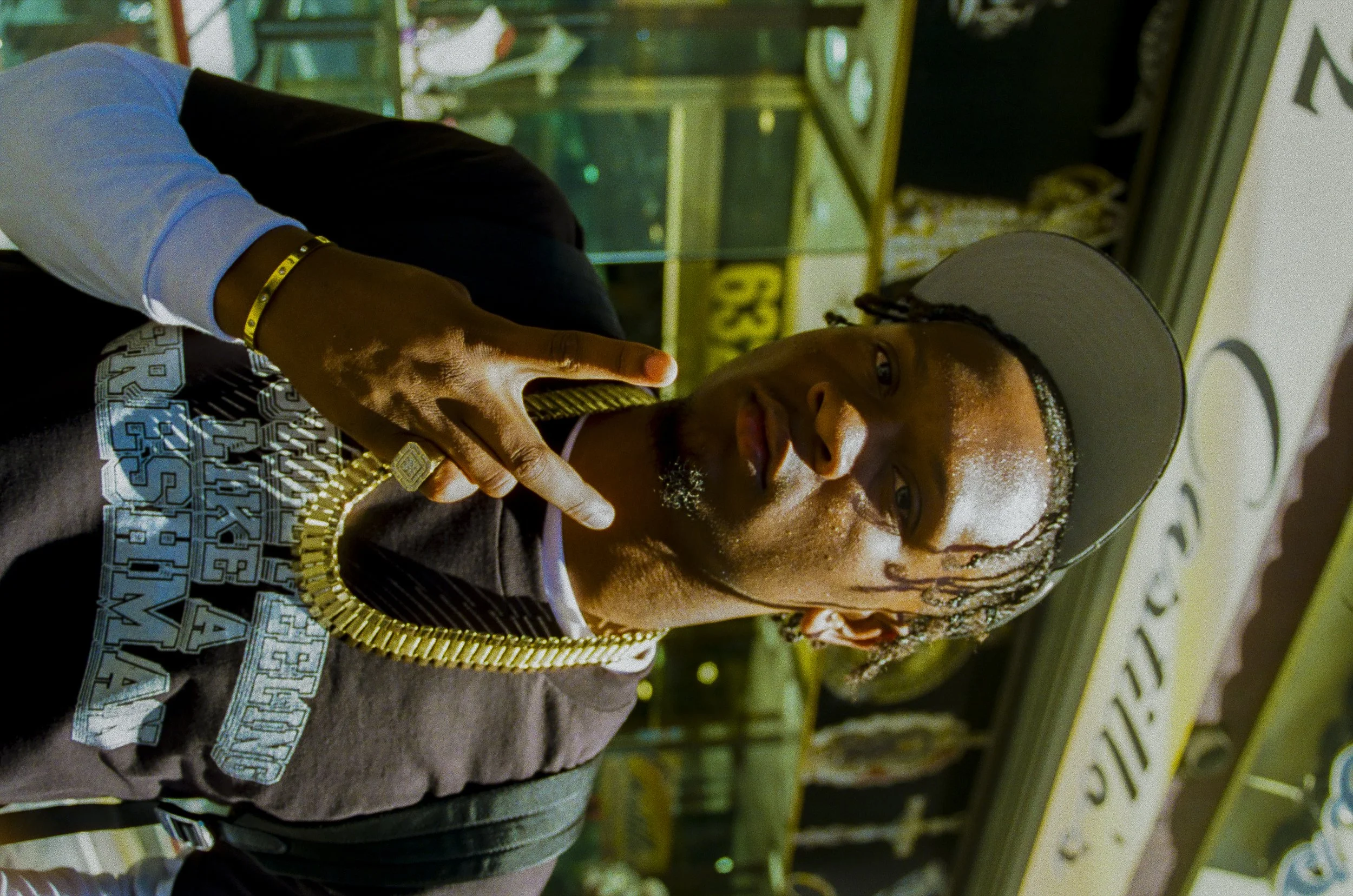 A young man with dreadlocks wearing a gray baseball cap, a black and gray hoodie, and gold jewelry, pointing his finger towards the camera, inside a store with shelves filled with various products.