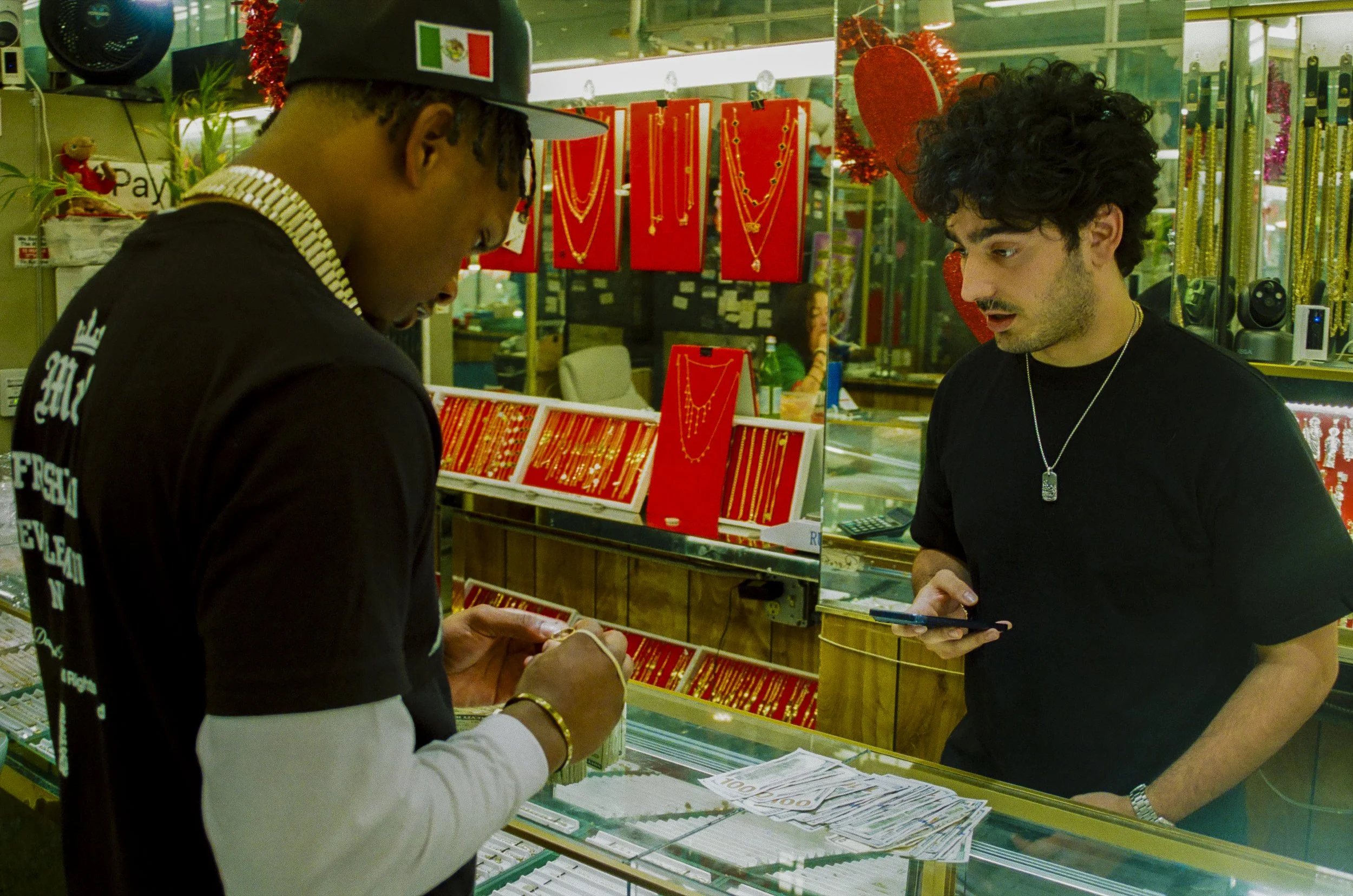 Two men at a jewelry store counter looking at jewelry. One man has dark curly hair and wears a black t-shirt, the other man has his head down and wears a hat with an Italian flag. The background shows red jewelry displays and gold jewelry.