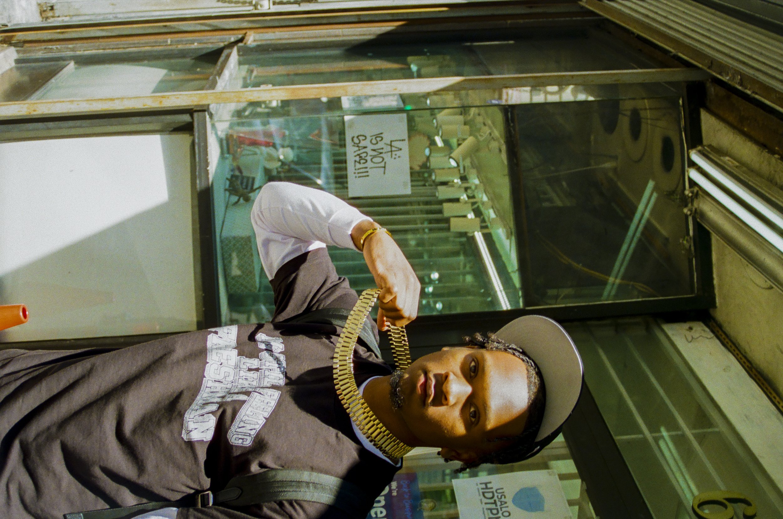 A man with a goatee and gold chain jewelry standing in front of a glass display case at a store, with other people and signs visible in the background.