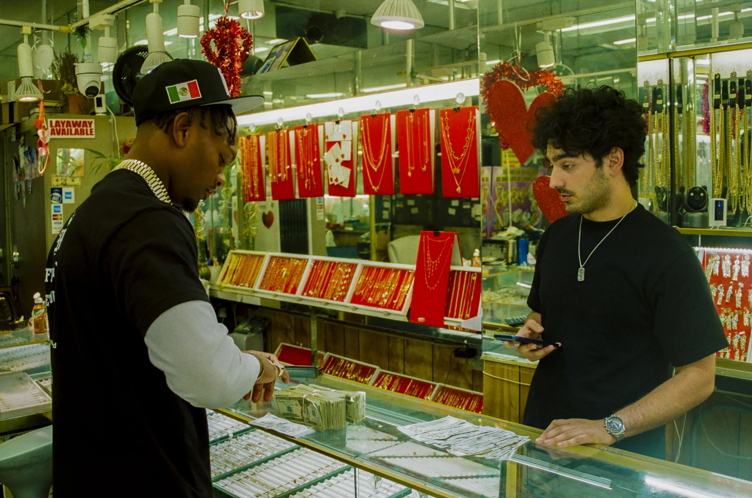 Two men are at a jewelry counter inside a store, examining jewelry displayed on red cards and a glass counter, with festive heart and tinsel decorations visible in the background.