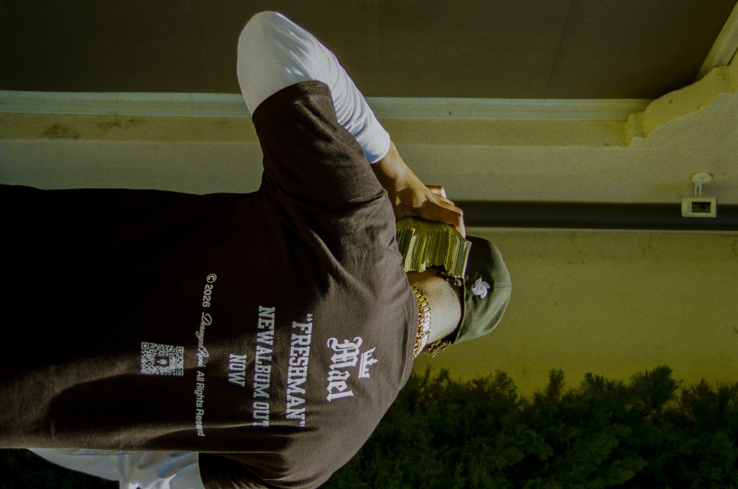 Person sitting outdoors at night, wearing a brown shirt with text and a QR code, holding a yellow object, with trees and power outlet in the background.