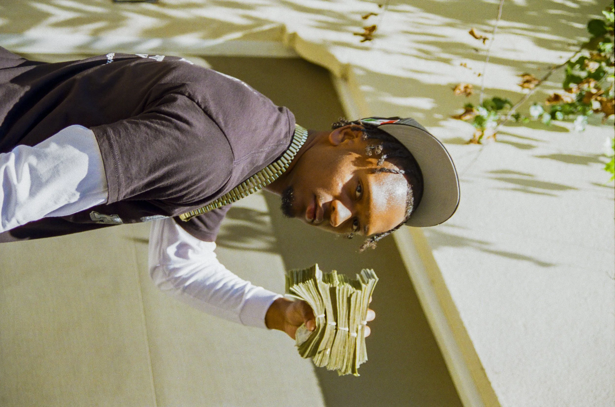 A man wearing a hat and a bejeweled necklace is leaning over a table with stacks of money, with a beige and green background.