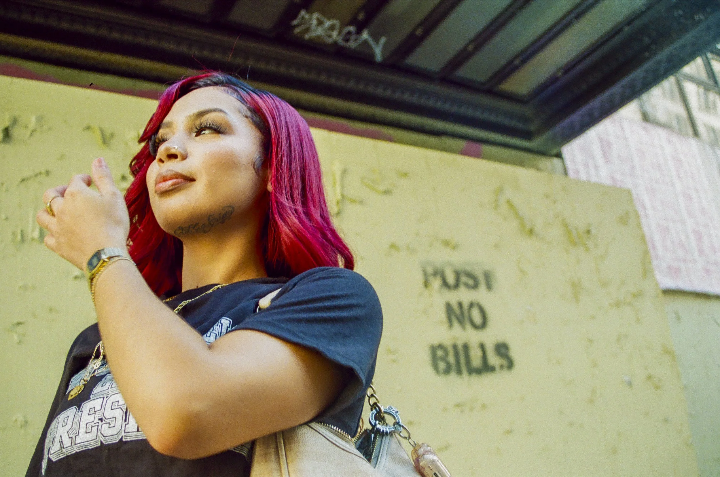 A woman with pink and black hair, wearing a nose ring, jewelry, and a dark blue shirt, standing outdoors near a yellow wall, with a street sign in the background.