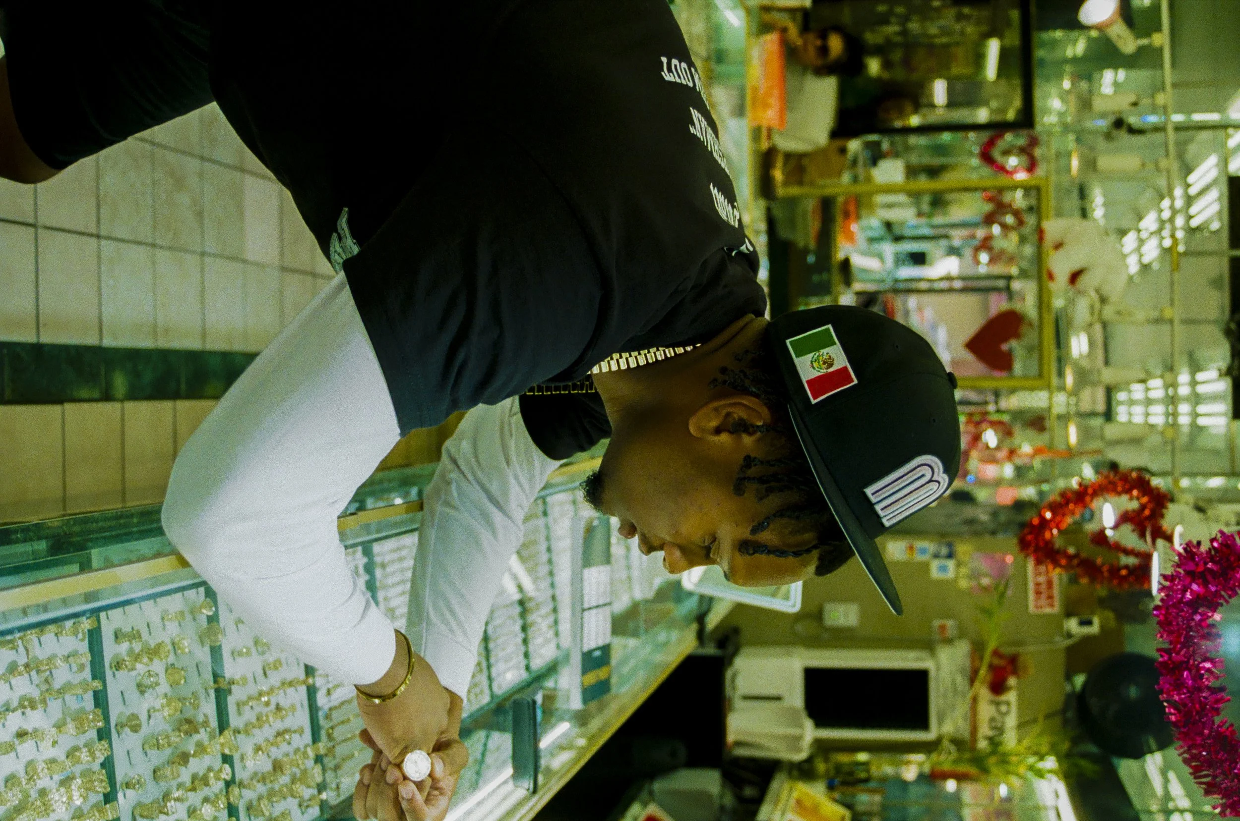 Two men are shopping for jewelry in a store, looking at display cases filled with rings and gold jewelry. One man is wearing a black hat with a Mexican flag and the other is in a white shirt.
