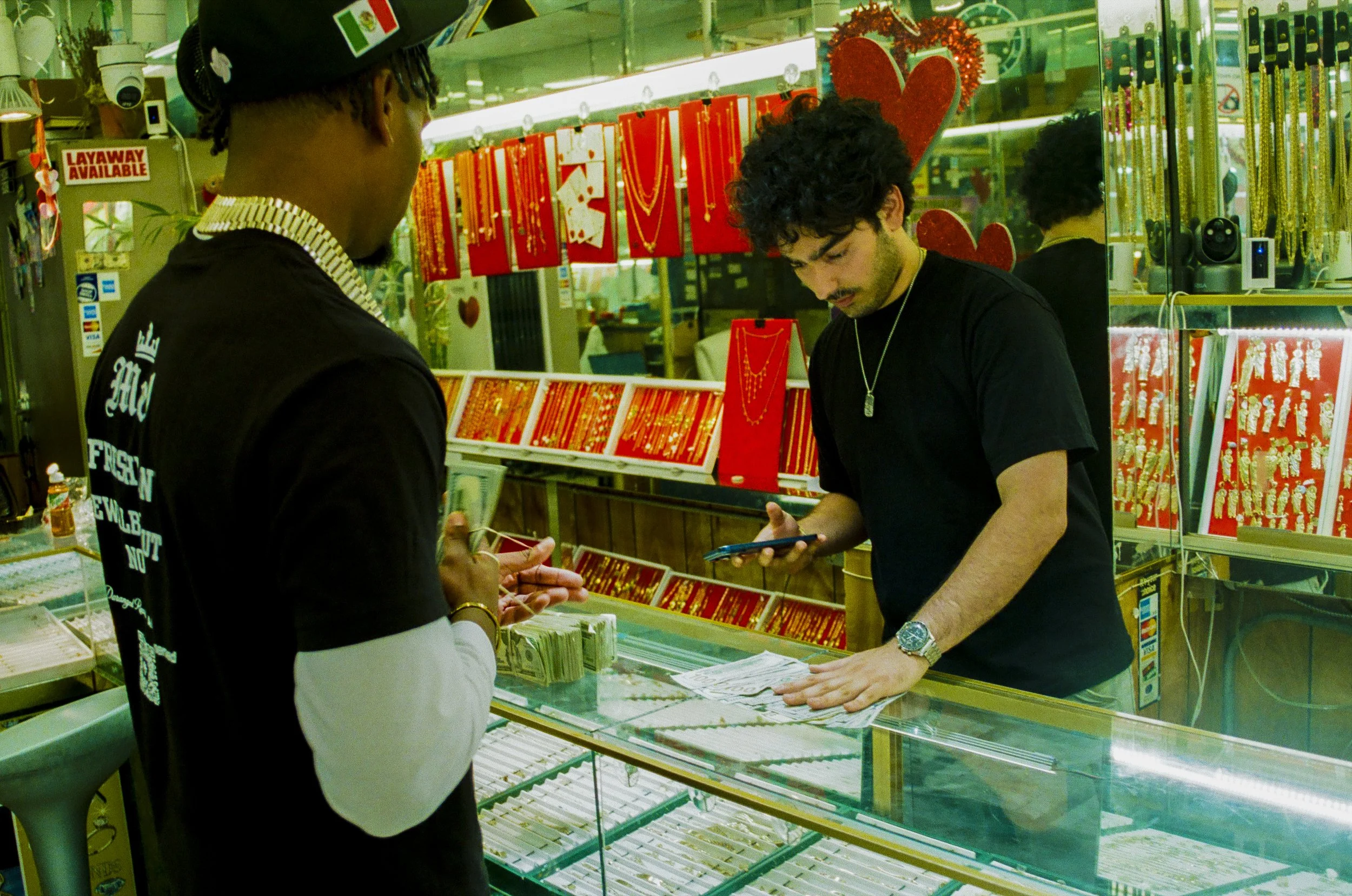 Two men standing at a jewelry store counter, one looking at jewelry and the other showing something on his phone. The background has red and gold jewelry displays and decorations.