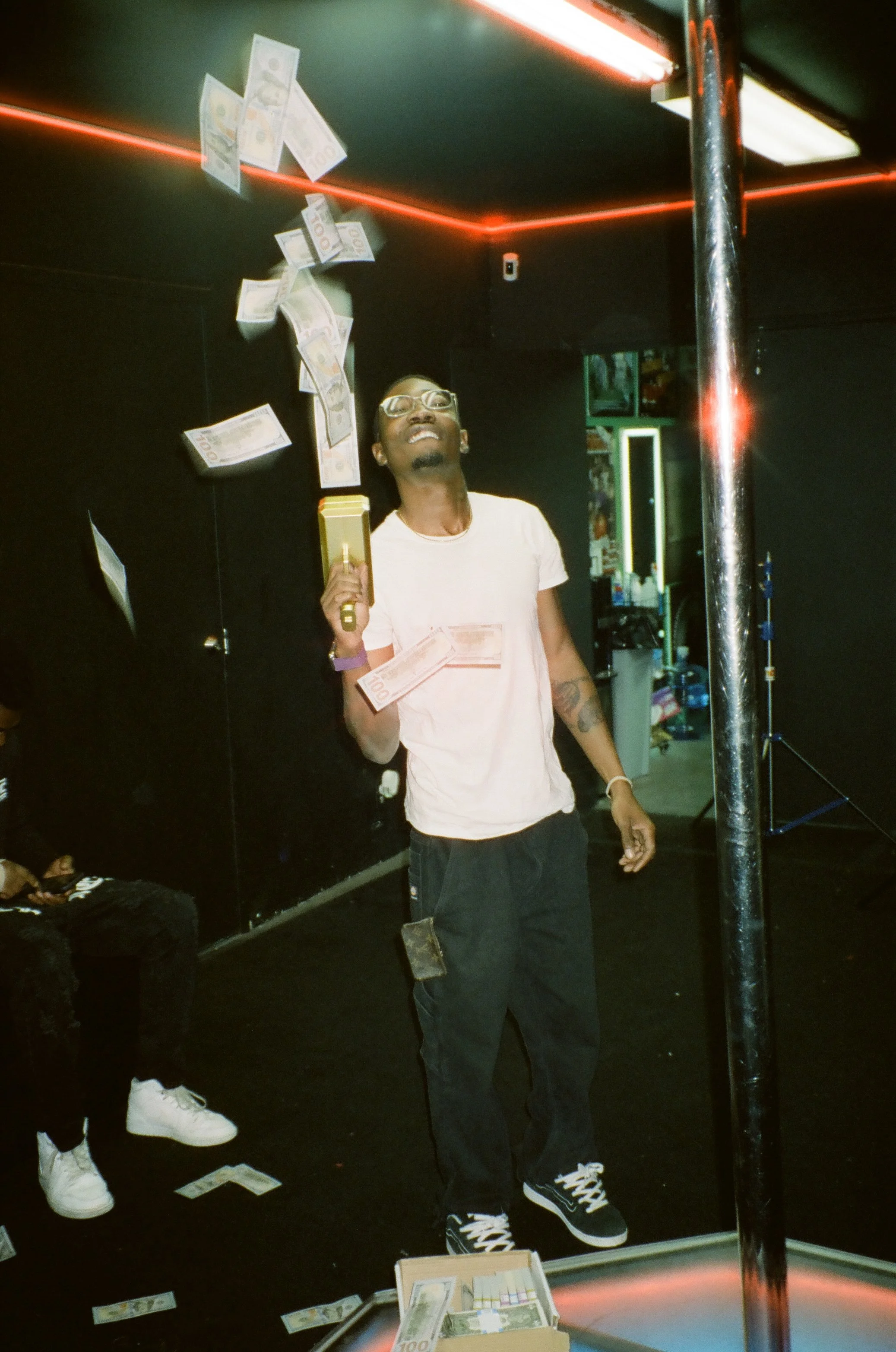 A man with glasses and tattoos on his arm is standing in a dark room, holding a stack of cash, with paper money floating around him. There is a box of cash on a table in front of him, and the scene appears to be inside a club or studio with neon lighting and a pole to the right.