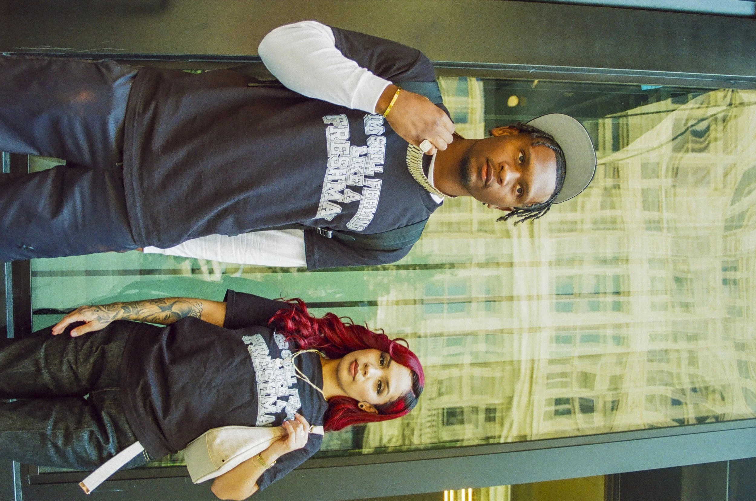 A man with braided hair, wearing a baseball cap and a graphic t-shirt, standing beside a woman with bright red hair and tattoos, inside a glass elevator or room with a patterned background.