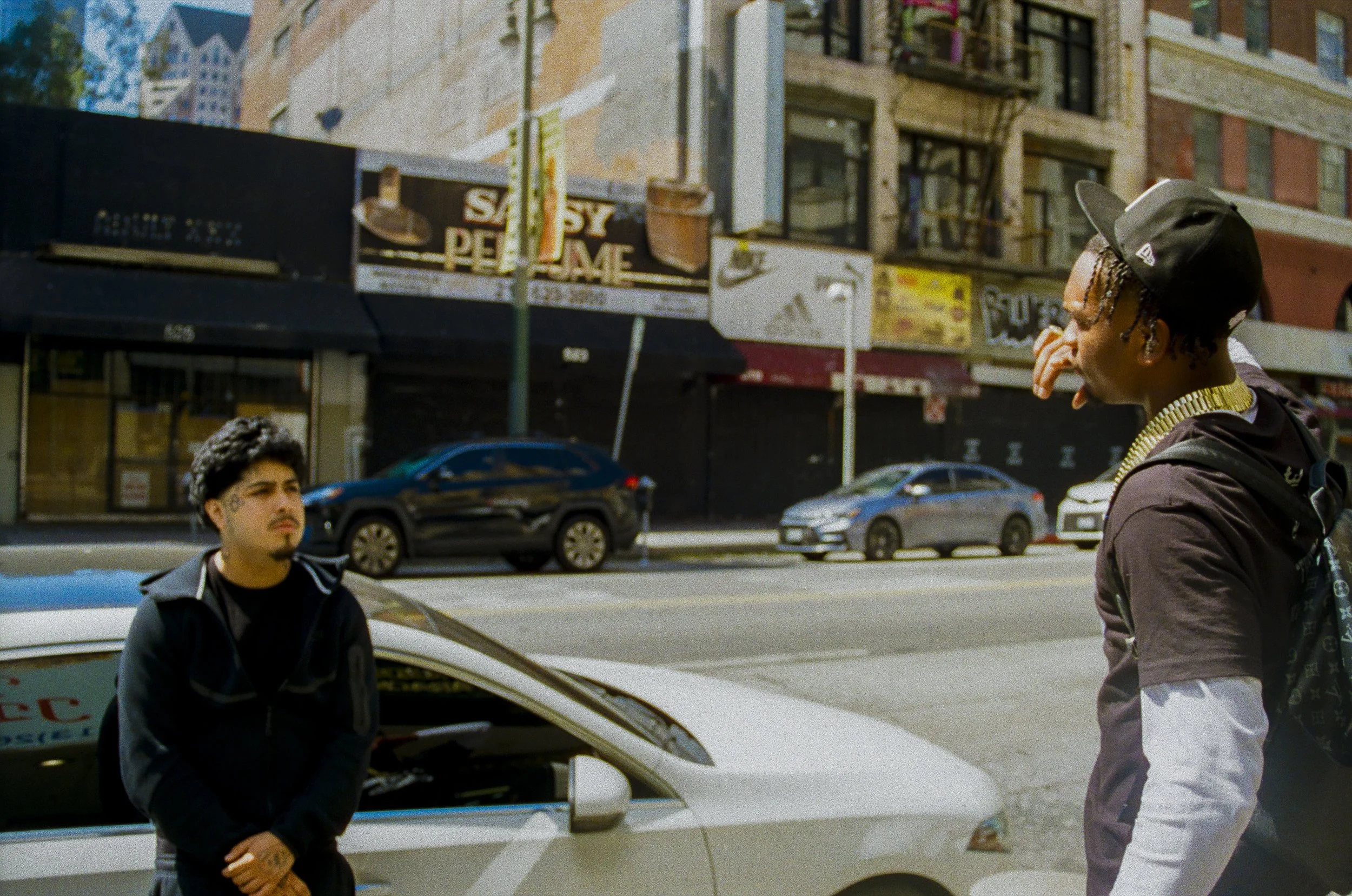 Two young men standing on the sidewalk in an urban area. One is wearing a black cap and jacket with a gold chain, facing sideways. The other has curly hair, wears a black jacket, and stands near a white car. Background shows retail storefronts, parke