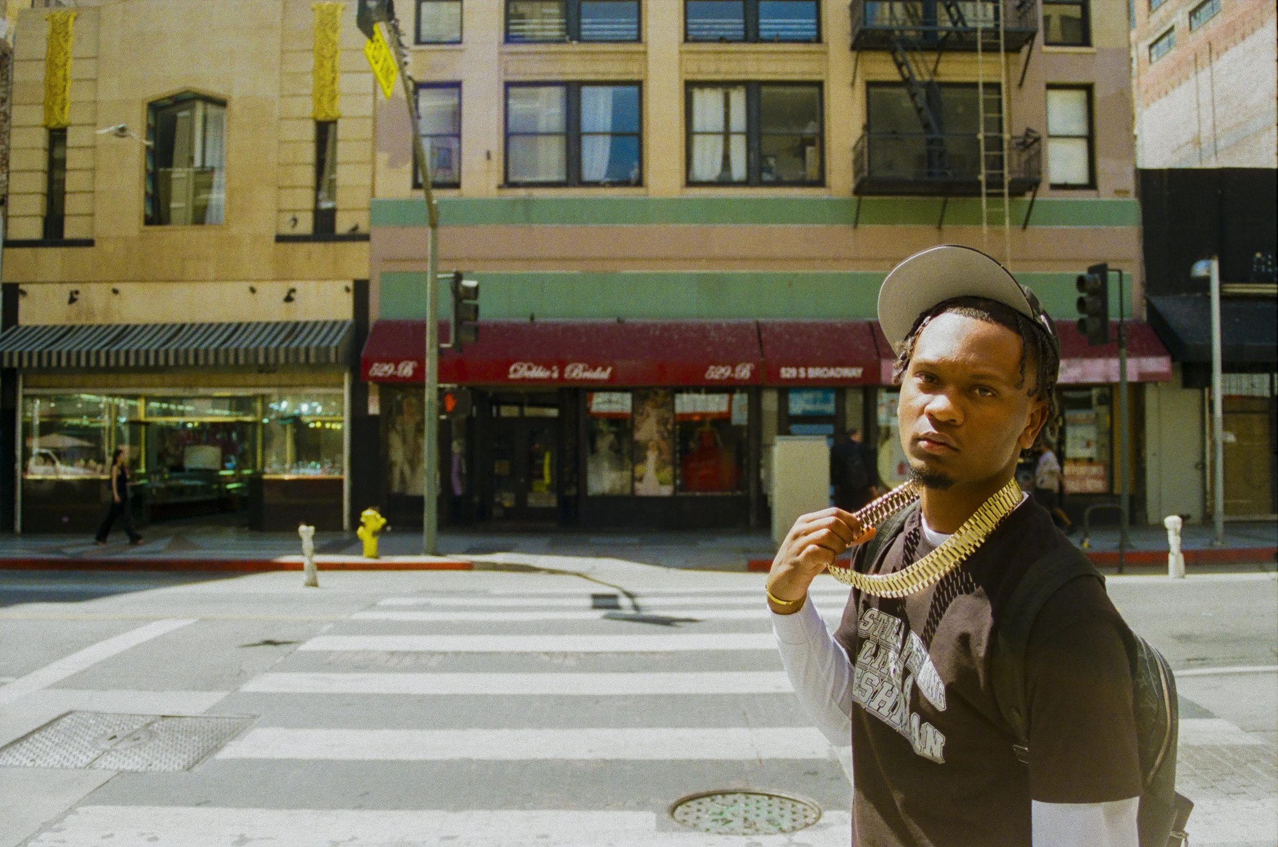Young man with braided hair wearing jewelry stands in front of city street background.