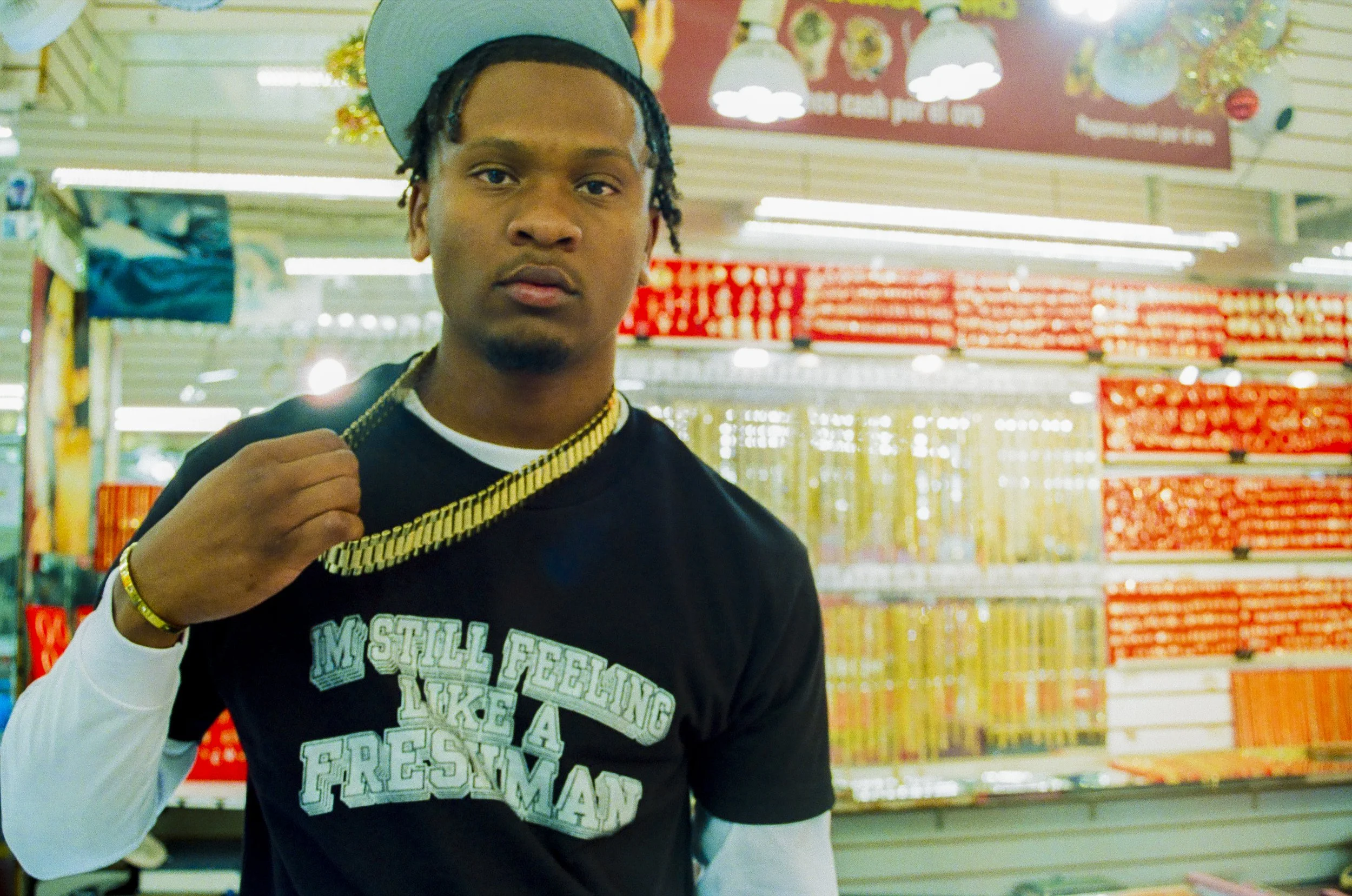 A young man standing in a grocery store aisle, looking at the camera while holding a gold chain around his neck. He is wearing a black T-shirt with white and teal lettering and a gray cap. The background shows shelves stocked with various products an