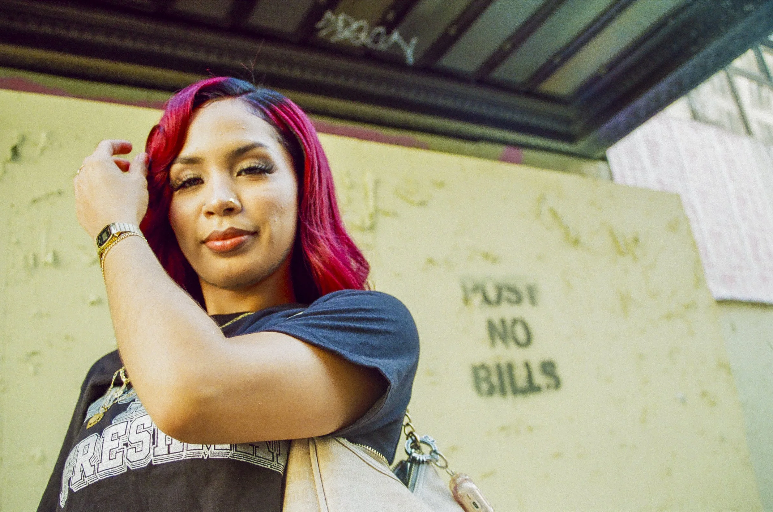 A woman with pink and purple hair, wearing a black T-shirt with white text, standing outdoors near a green wall with faded writing. She has a watch, rings, earrings, and a chain on her wrist, with a necklace, and is touching her hair while smiling.