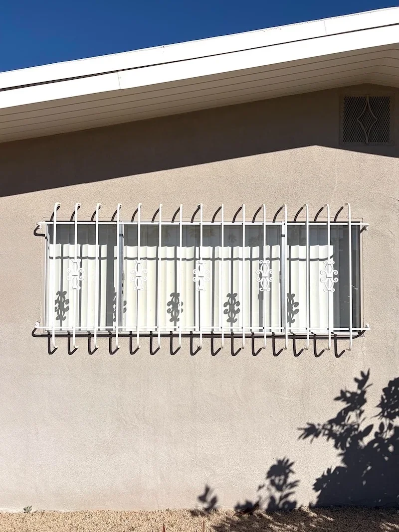 white window grilles with decorative scrolls covering the windows of a tan home with the shadow of a plant upon the wall