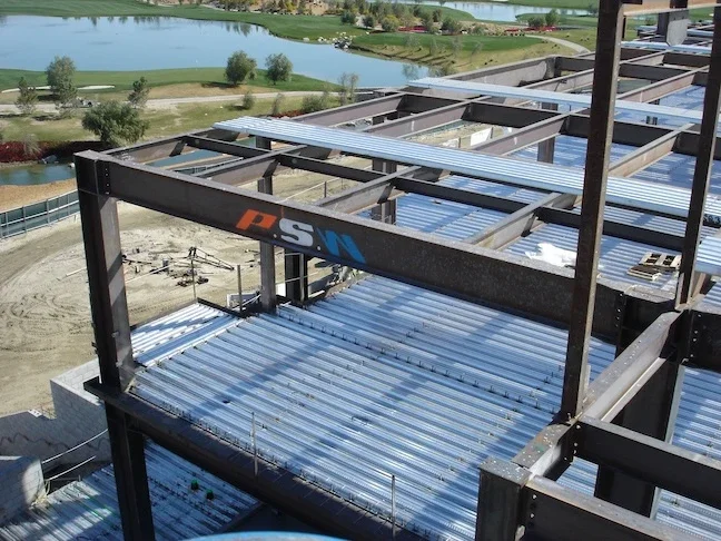 structural steel foundation for a building with beams and corrugated metal with the letters P.S.W. written in red, white, and blue on the front beam and a golf course with a lake in the background