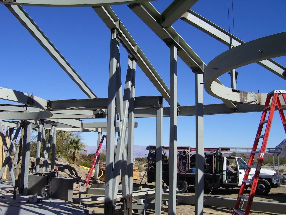 structural steel base for a complex building with circular rolled beams, sloped roof beams, and columns in a desert setting with a work car behind