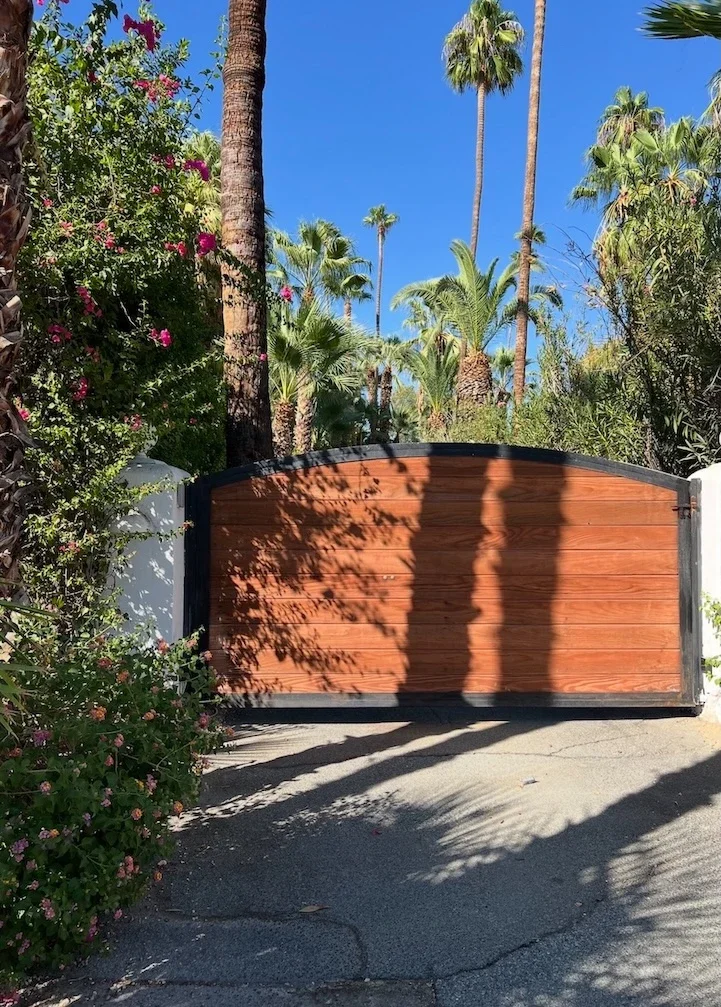 asphalt leading into a red-hued wood-paneled swing entry gate with a black steel frame attached to white pillars with palm trees surrounding