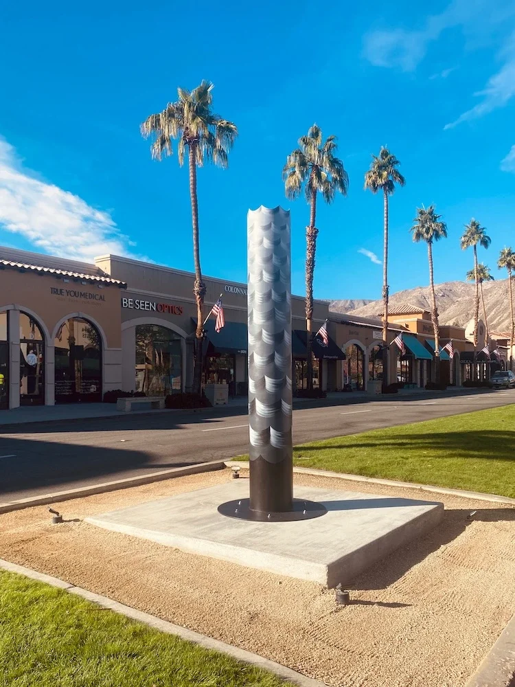 steel pole sculpture with black and grey painted waves in the median of a shop-lined street