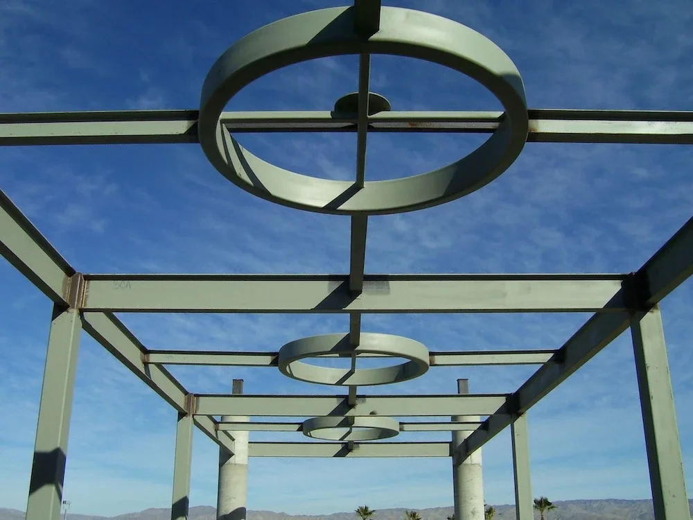 green steel beams and columns that form a walkway with circular rolled beams lining the structure’s center and blue sky behind