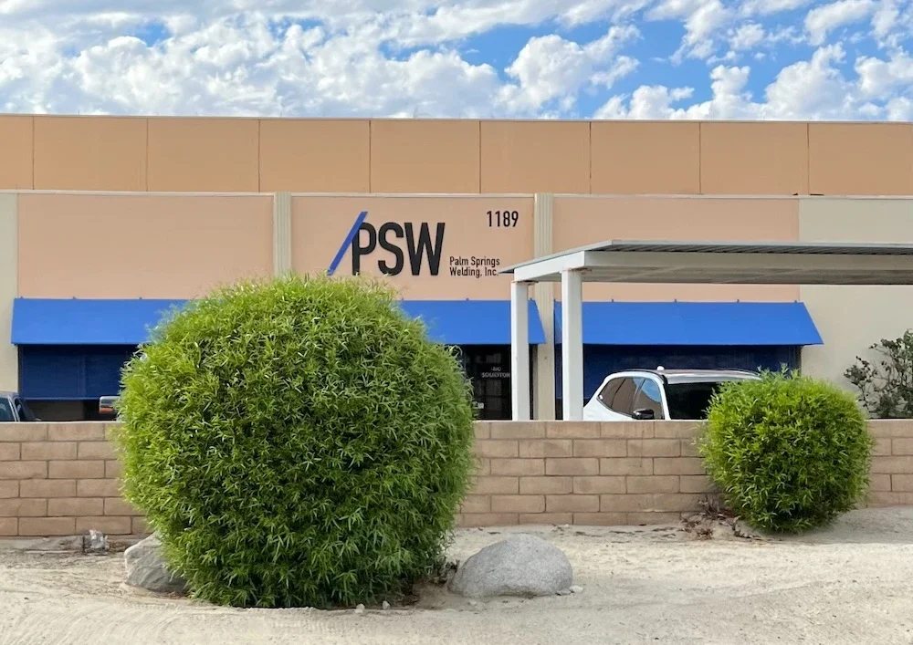 street view of a tan building with PSW, Palm Springs Welding, Inc., and 1189 written on it, blue awnings, a small carport with a car underneath, and a bush in front of a wall