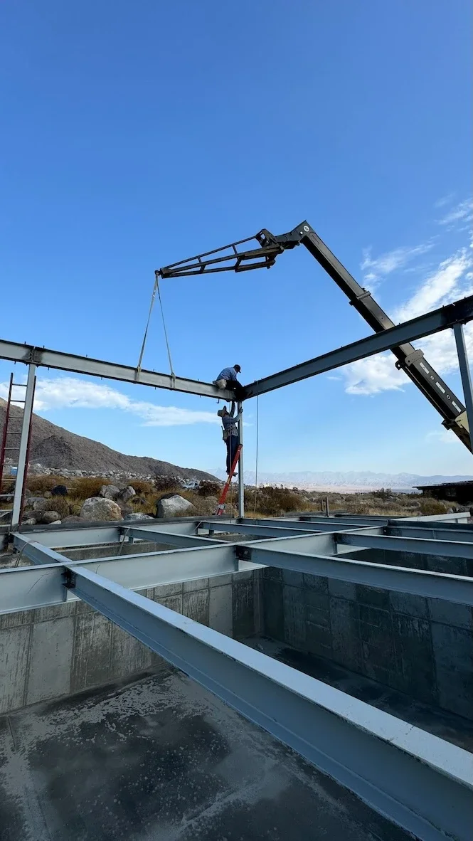 two men installing structural steel with a crane assisting for a structure with floor beams in a desert setting