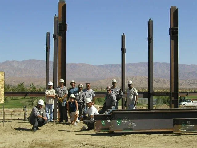 early-to-mid-2000s photo of a group of people with construction hats on next to the structural steel base for a building