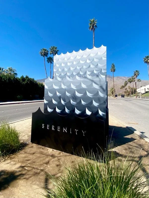 sculpture with black and grey painted waves on three layered steel sheets with the word “serenity” cut out on the bottom layer located in a street’s median