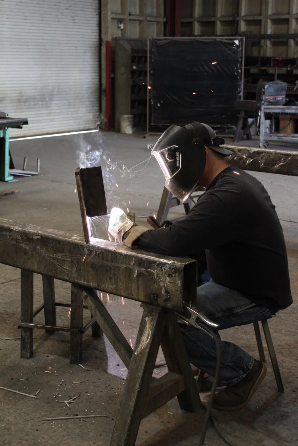seated welder with protective mask and gloves welding in a shop with sparks flying