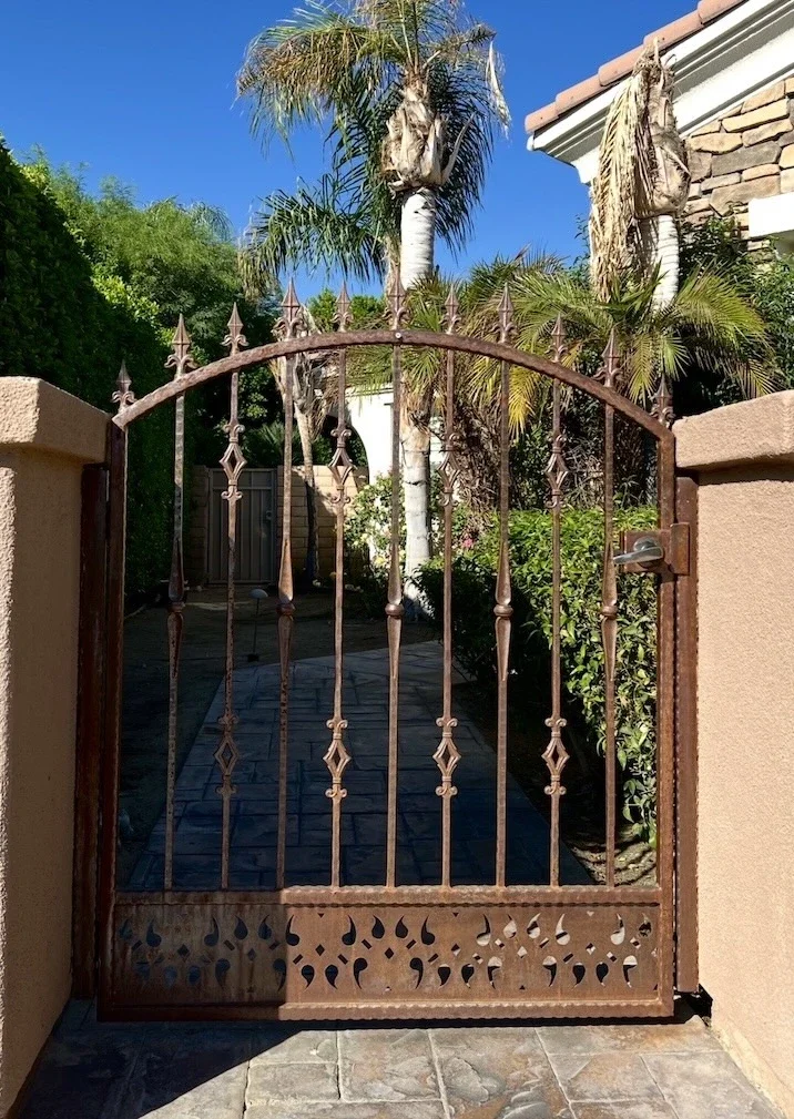 ornamental front gate with diamond hammered balusters, a teardrop cut-out base, and diamond pickets in a rust finish with a gold handle attached to brown pillars in the courtyard of a home