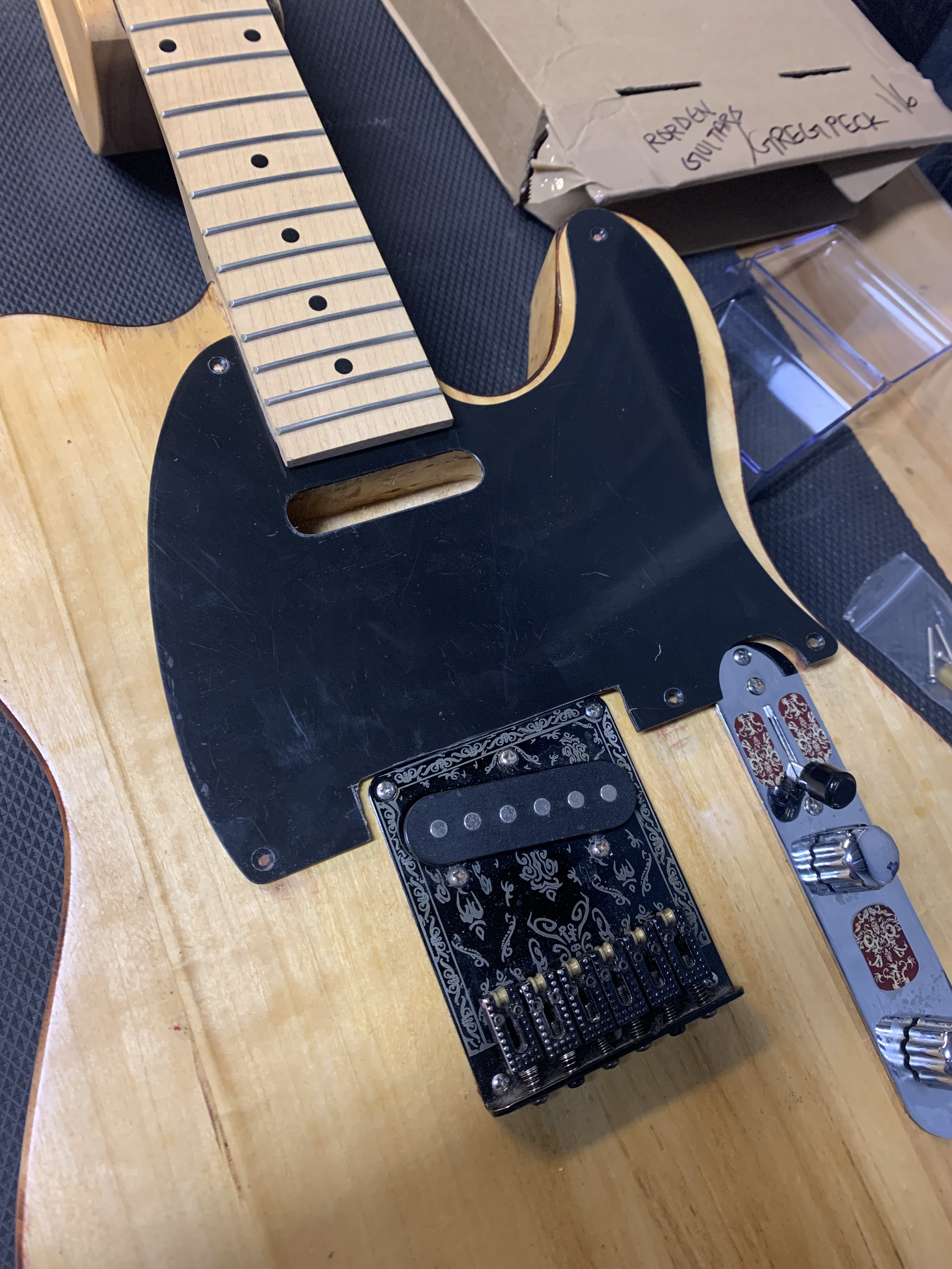 Partially assembled electric guitar with a wooden body, black pickguard, maple neck with frets, and decorated pickup and bridge. Cardboard boxes in the background.