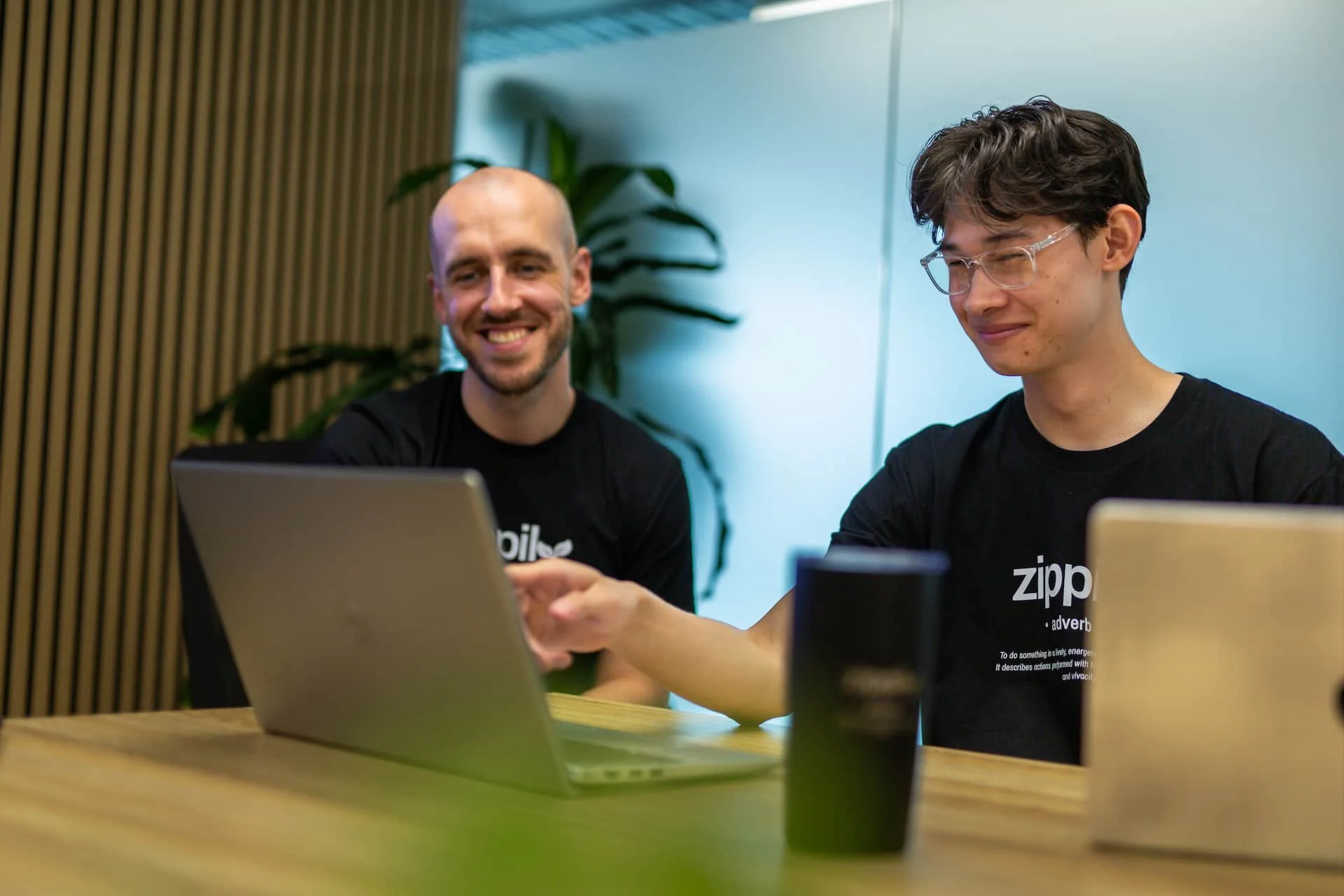 Two Auckland based HubSpot Specialists sitting at a wooden table with laptops, smiling and engaging in conversation. One is bald with a beard, the other has wavy dark hair and glasses. There is a large plant in the background.