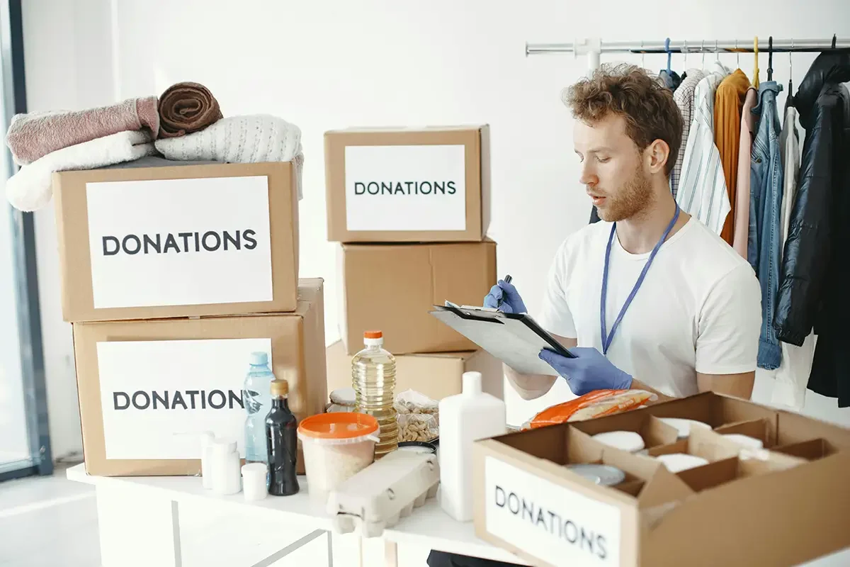 A man in a white shirt wearing blue gloves is writing on a clipboard at a table filled with donation items, including boxes labeled 'DONATIONS,' bottles, canned goods, and folded clothes in the background.