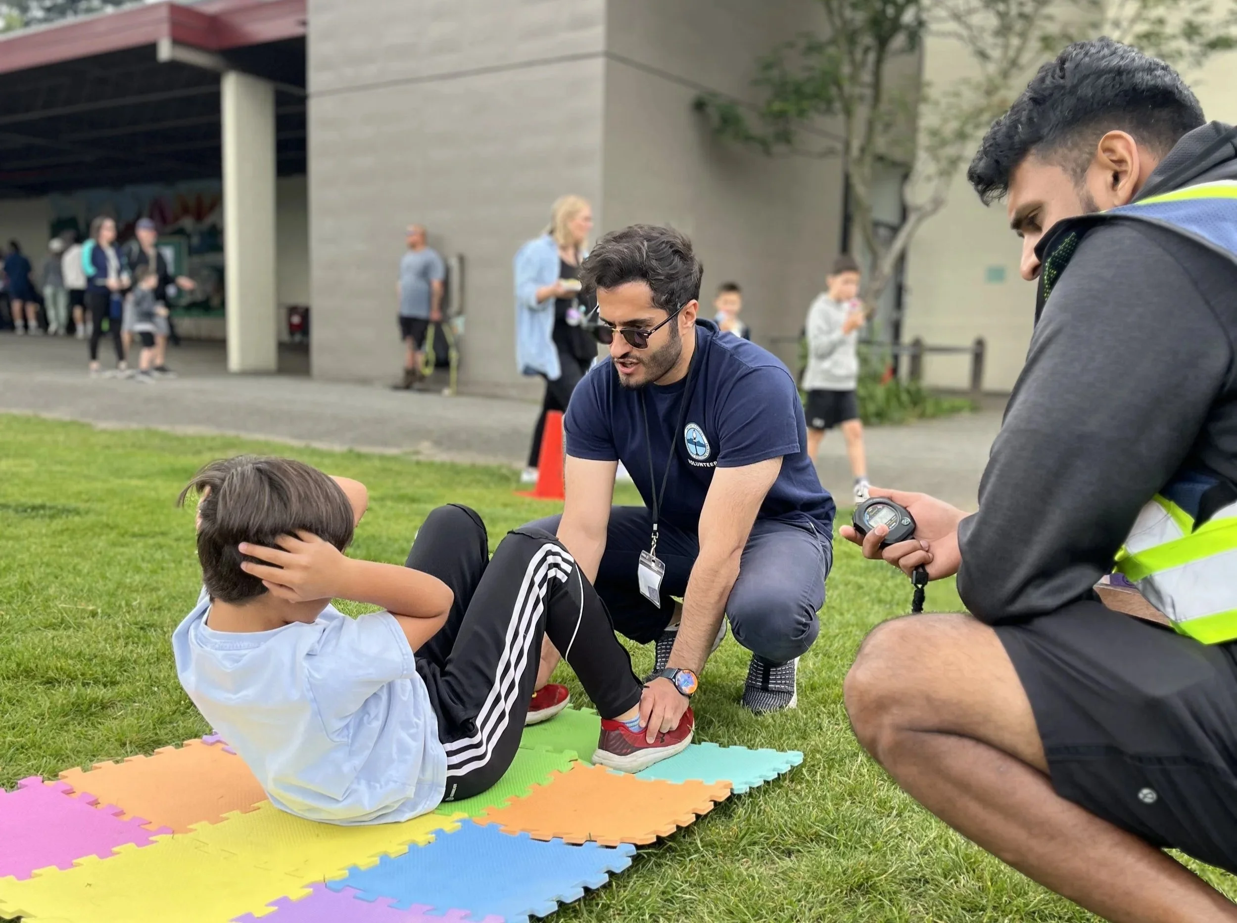 A young boy doing sit-ups on a colorful foam mat outdoors, being assisted by a man in sunglasses and a navy blue shirt. A person in a gray hoodie with a safety vest and a stopwatch kneels nearby, recording or timing the activity. Several people are in the background, some standing and some walking, near a building with a covered area and trees.