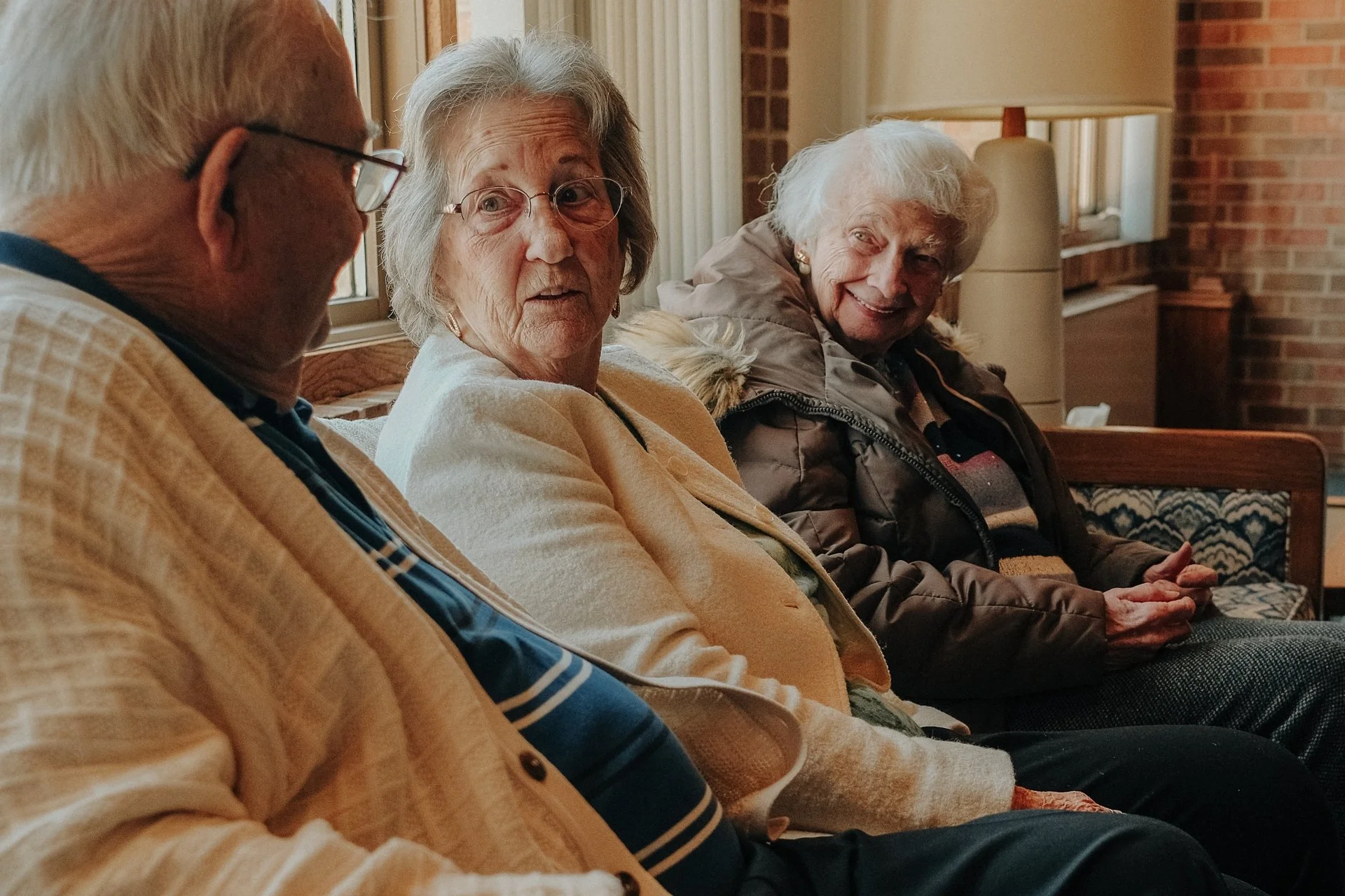 Three elderly people sitting on a couch in a cozy room, engaging in conversation and smiling.