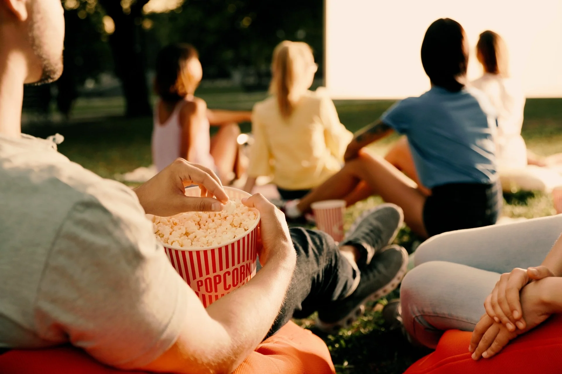 People sitting on the grass watching a movie outdoors at sunset, with one person holding a bucket of popcorn.