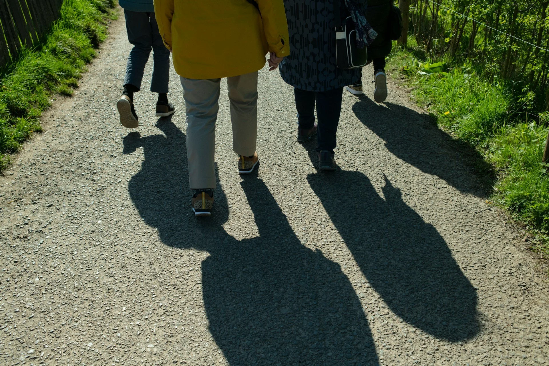People walking on a gravel path with backpacks, side shadows visible on the ground, greenery on the sides.