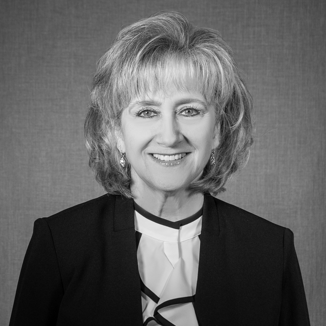 Black and white professional headshot of a smiling woman with shoulder-length hair, wearing a dark blazer and a patterned blouse, against a plain background.