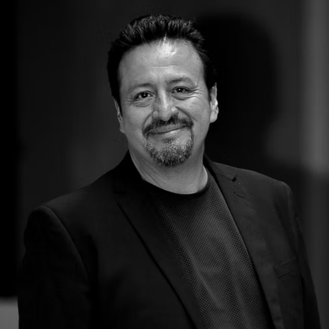 Black and white portrait of a man smiling, wearing a blazer and a patterned shirt, with dark background.