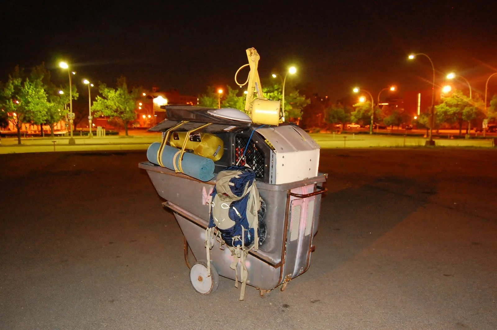 A metal cart filled with camping gear, including a blue sleeping mat, yellow bag, and backpack, parked on a night street with trees and streetlights in the background.