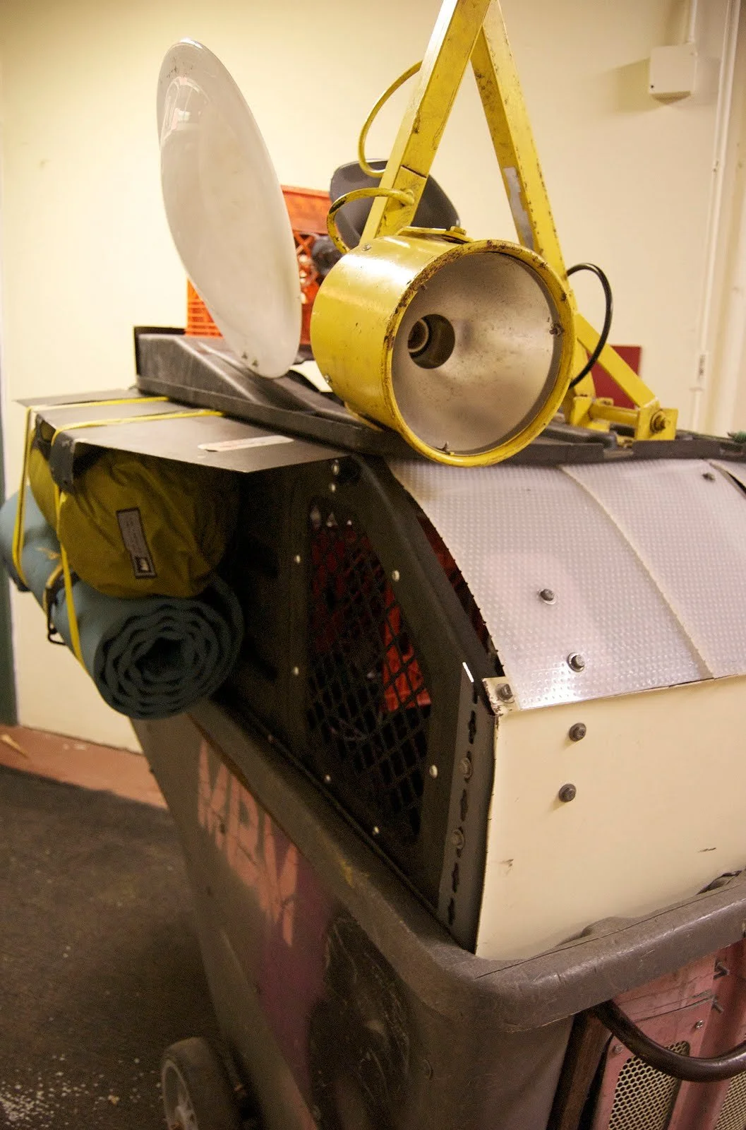 A metal cart with various equipment, including a yellow portable floodlight, a rolled-up camping mat, and a dish reflector, stored on a black shelf and on top of the cart.